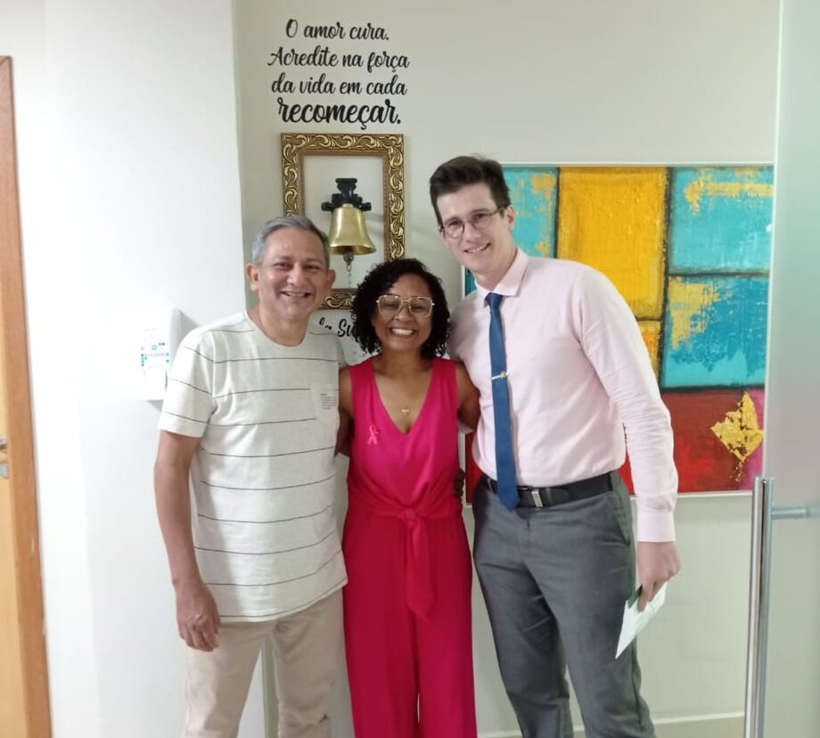 O marido de Risélia, Denilson, Risélia e o médico responsável por ela, Dr. Lucas Vian, diante do sino da cura.