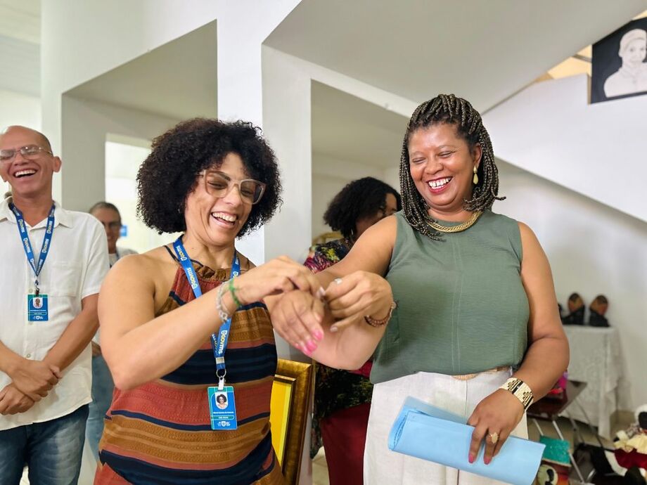 Técnica da Subsecretaria de Políticas Públicas para Promoção da Igualdade Racial, Márcia Catarina colocando a fitinha Novembro Negro, MS sem Racismo no braço da subsecretária Vânia