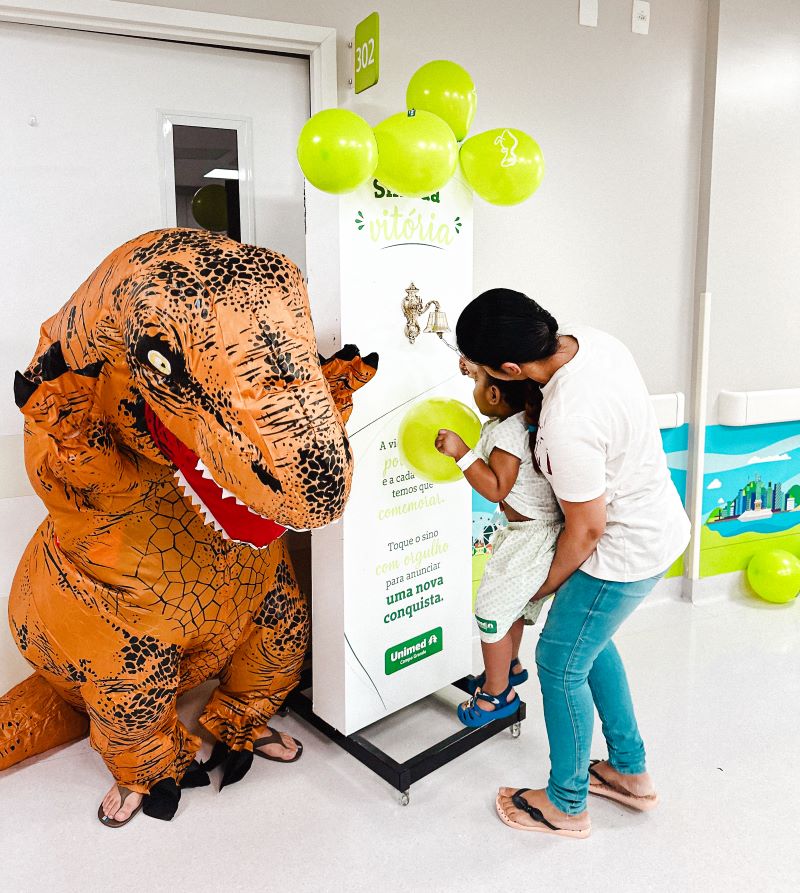Ao lado da mãe, Davizinho toca o sino da vitória sob o olhar atento do dinossauro e da equipe, marcando o fim de uma etapa difícil com leveza e emoção.