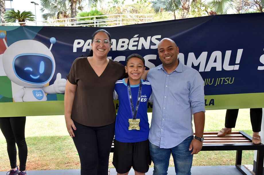 Lucas Jamal ao lado dos pais, comemorando a medalha de ouro conquistada no World Jiu-Jitsu Championship, com faixa de homenagem ao fundo.