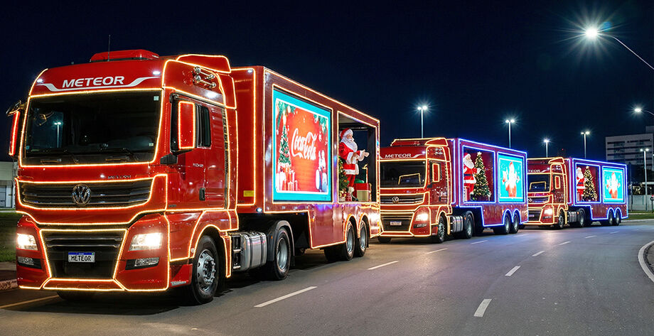 Caminhões iluminados da Caravana de Natal desfilando à noite, com público assistindo nas calçadas.