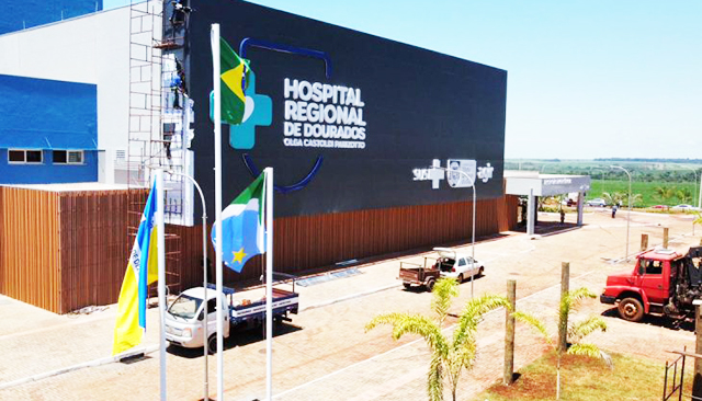 Fachada do Hospital Regional de Dourados Olga Castoldi Parizotto, às margens da BR-463, com entrada principal, letreiro e equipe de saúde na área externa.