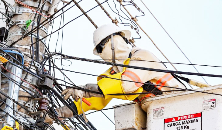 Poste urbano com técnicos desmontando fiação e removendo cabos com rua de centro ao fundo.