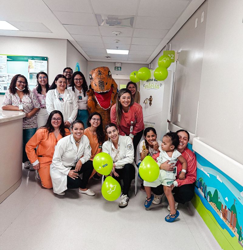 Equipe do Hospital Unimed Campo Grande celebra a alta de Davizinho com balões, sorrisos e um dinossauro especial, símbolo de carinho e superação.