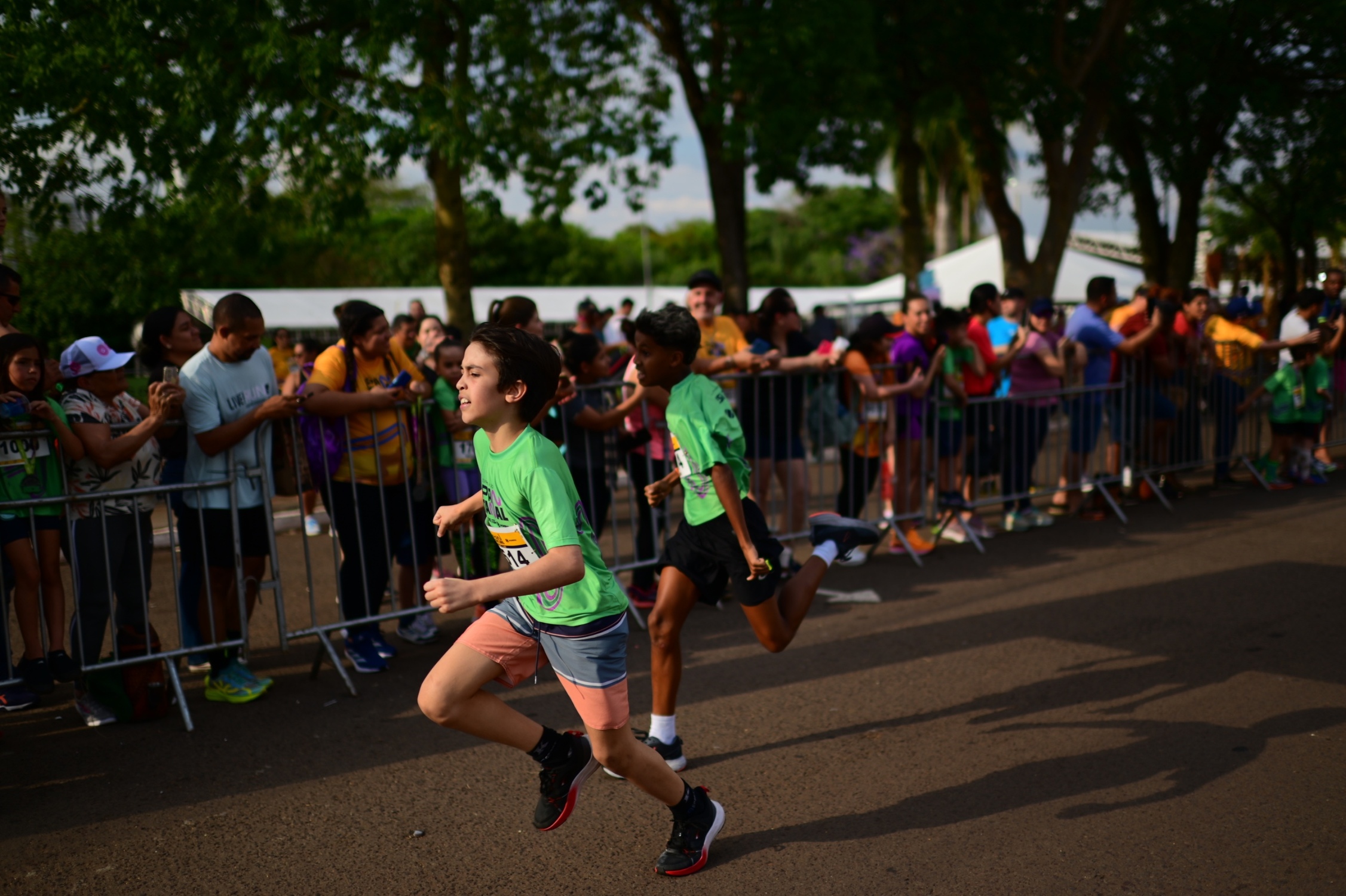 Garotos mostram energia e concentração durante a corrida, enquanto o público acompanha e apoia ao longo do trajeto da Corrida Kids.