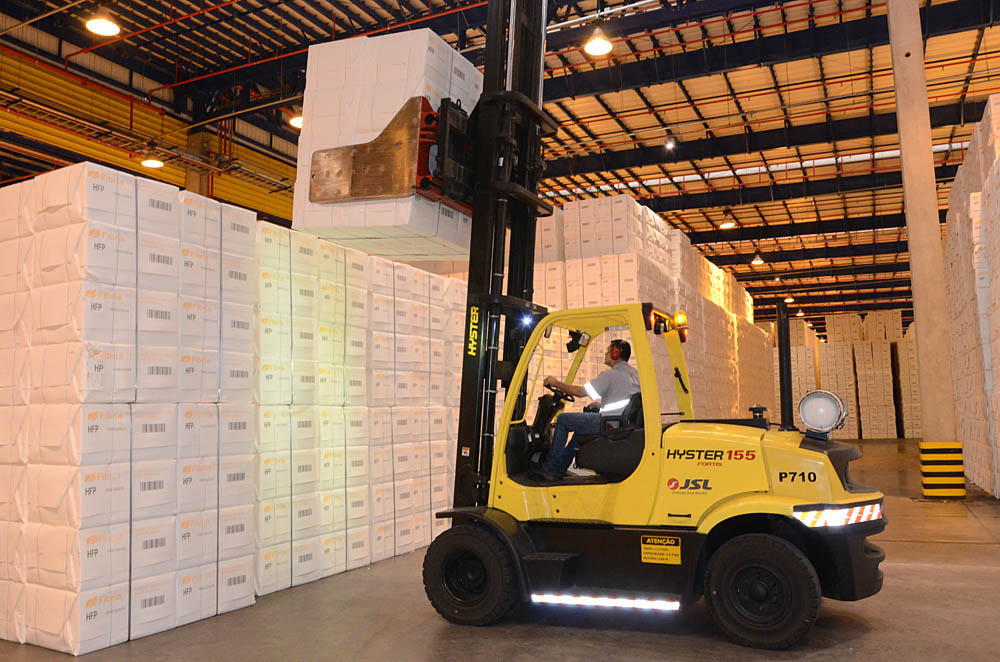 Operador movimenta fardos de celulose com empilhadeira em centro logístico, evidenciando a cadeia de armazenamento e distribuição da produção industrial.