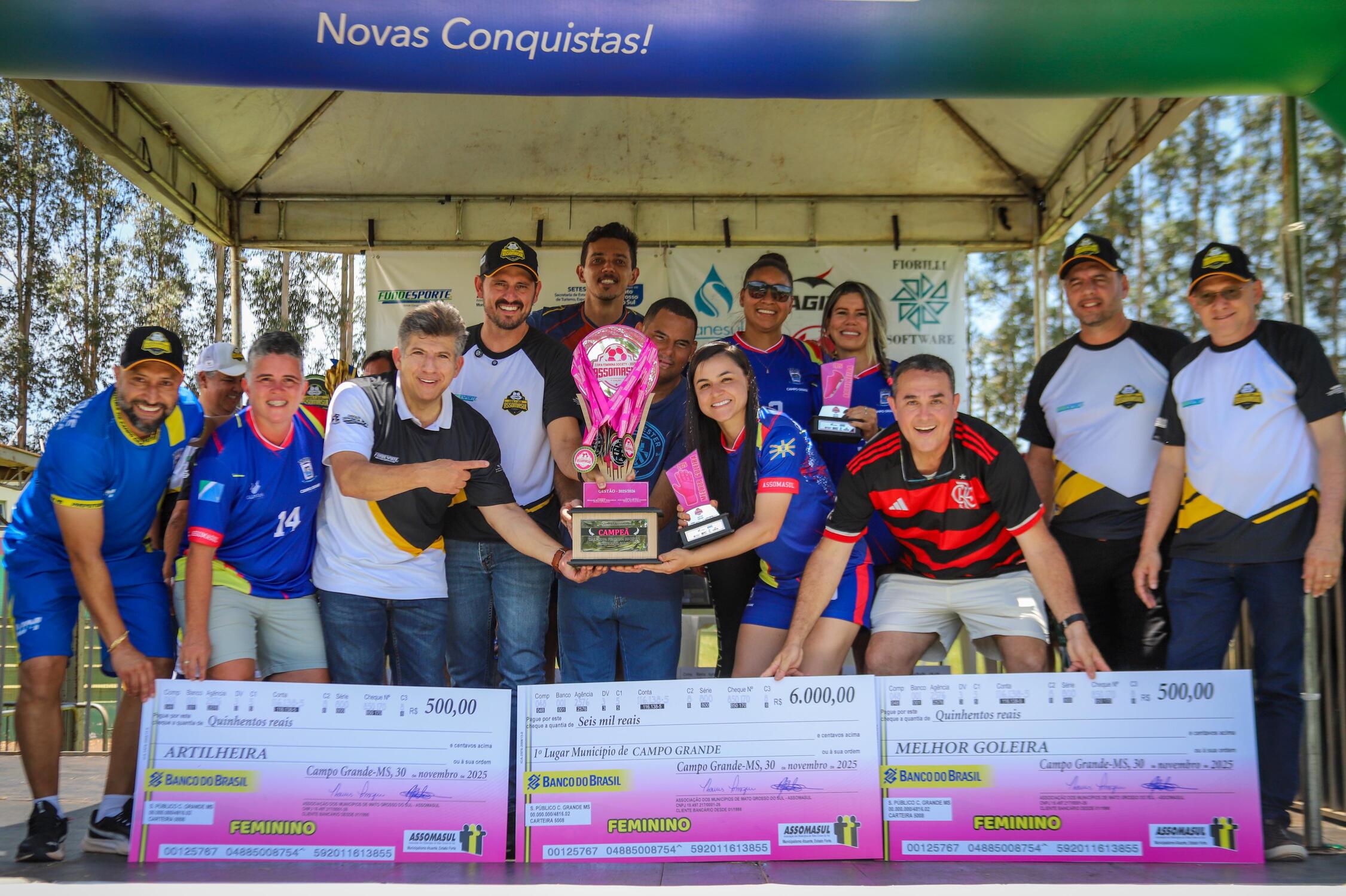 Equipe feminina de Campo Grande comemora o título da Copa Assomasul 2025 ao lado de dirigentes, com troféu em mãos e cheques de premiação em destaque.